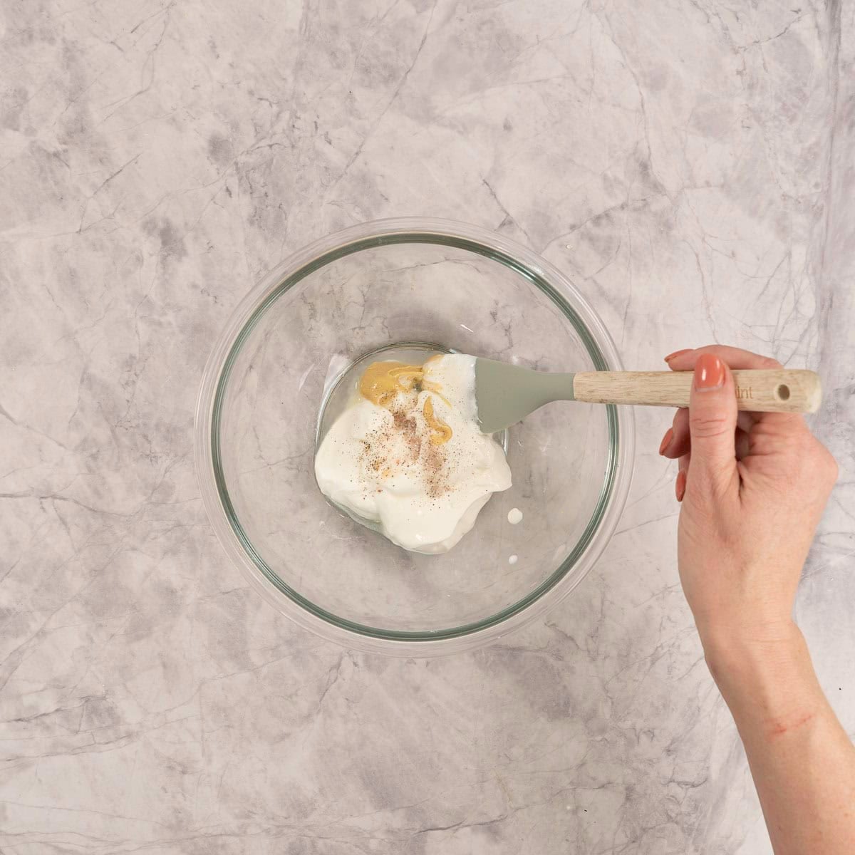 Large glass bowl on marble bench top with ingredients for dressing inside, hand holding a spatula stirring.