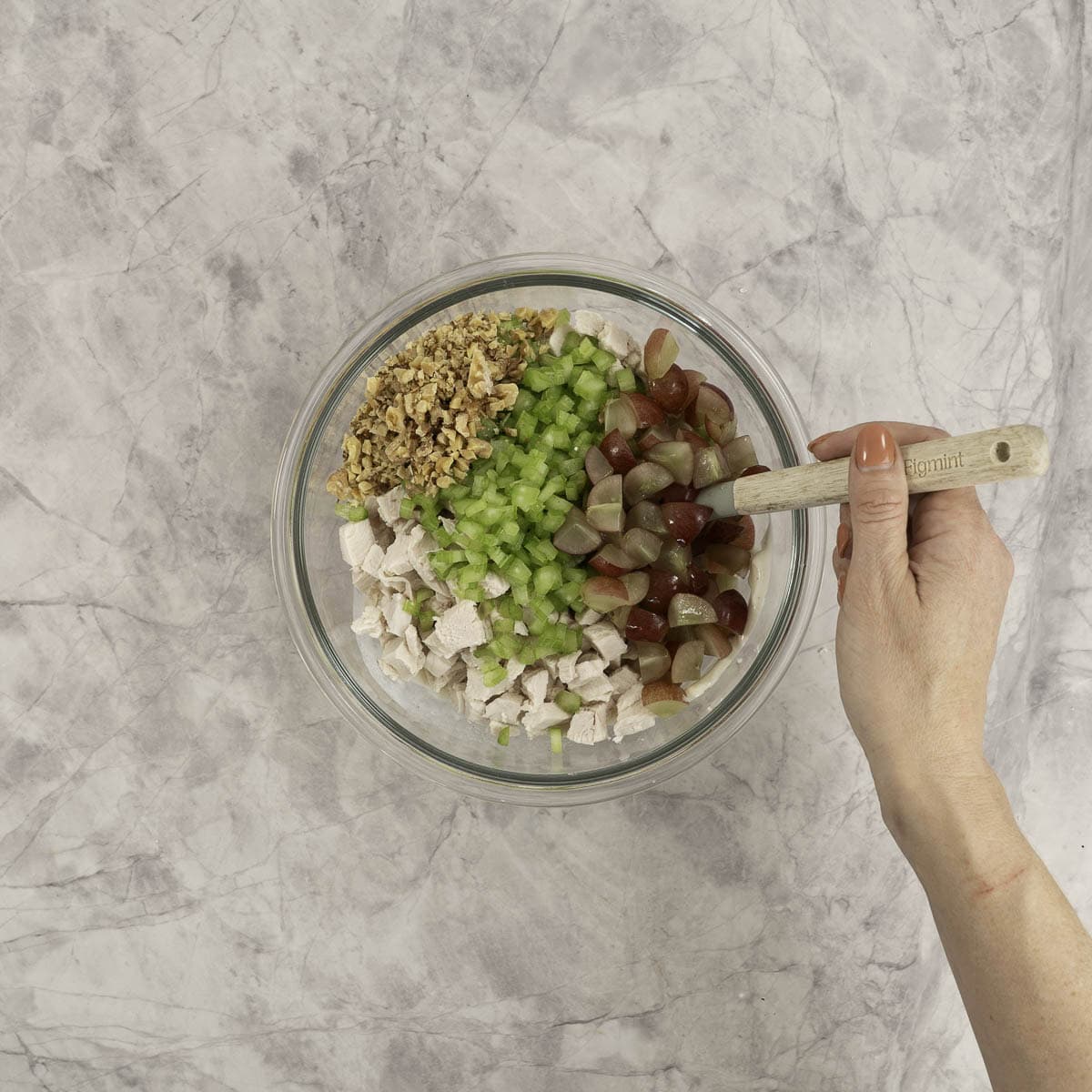 Chopped up celery, chicken, grapes and walnuts added into sauce mixture in large glass bowl on marble benchtop with woman's hand holding a spatula.
