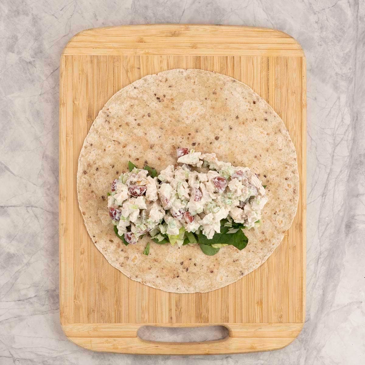 Wooden chopping board with wrap and salad chicken filling to one side sitting on a bed of lettuce.