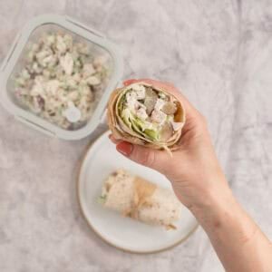 Woman's hand holding a half of a wrap showing the filling, container with extra salad and plate with other half of wrap on bench top below.