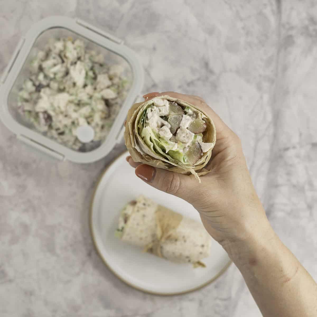 Woman's hand holding a half of a wrap showing the filling, container with extra salad and plate with other half of wrap on bench top below.
