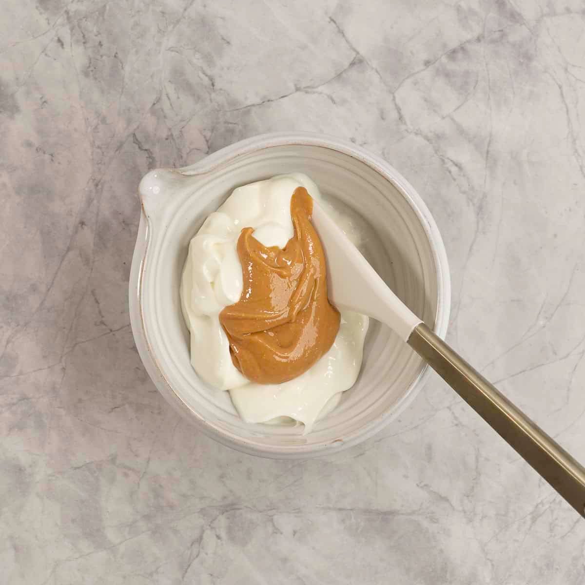 Yogurt and peanut butter in a white bowl on a marble bench top with spatula inside.