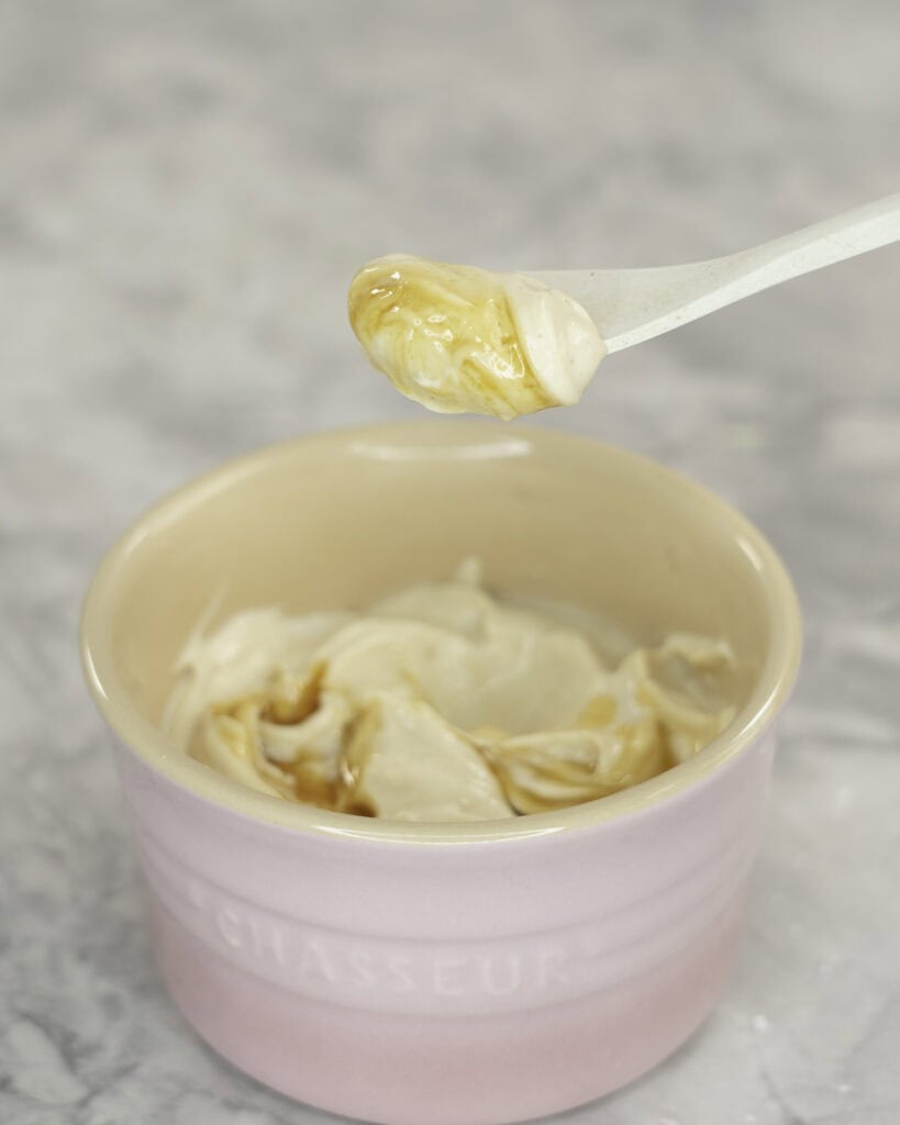 Creamy mixture with maple syrup drizzled on top in a small blush pink ramekin, white spoon taking a scoop out on marble bench top.
