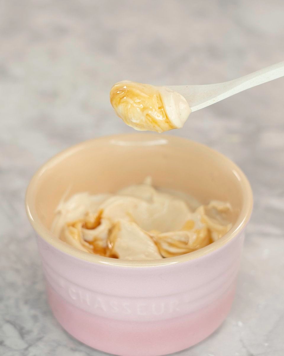 Creamy mixture with maple syrup drizzled on top in a small blush pink ramekin, white spoon taking a scoop out on marble bench top.