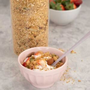 Small blush pink bowl with granola, yogurt and a spoon inside with a large glass jar of granola in the background.