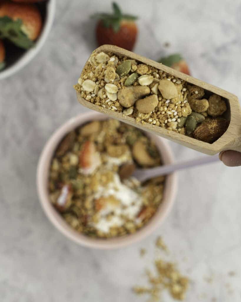 Small wooden scoop with granola inside held above a blush pink bowl with granola, yogurt and a spoon inside.