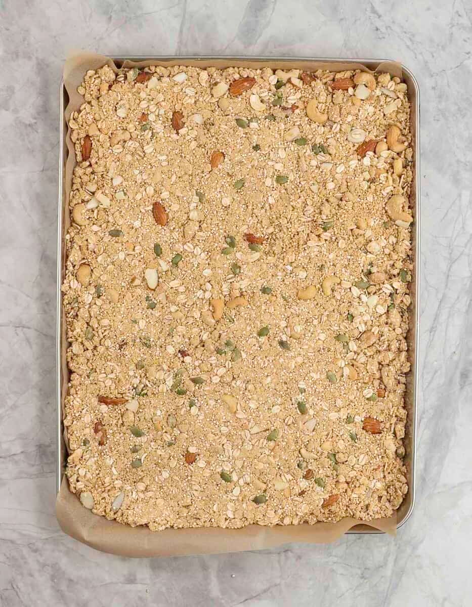 Raw granola thinned out on a lined baking tray on marble bench top.