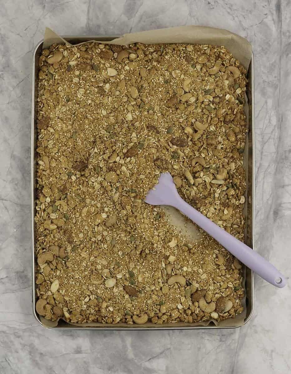 Toasted granola thinned out on a lined baking tray with purple spatula inside.
