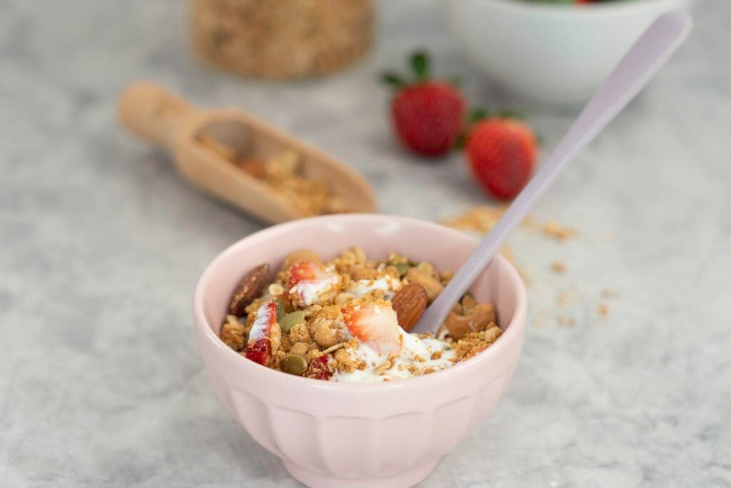Small blush pink bowl with granola, yogurt and a spoon inside with a large glass jar of granola in the background.