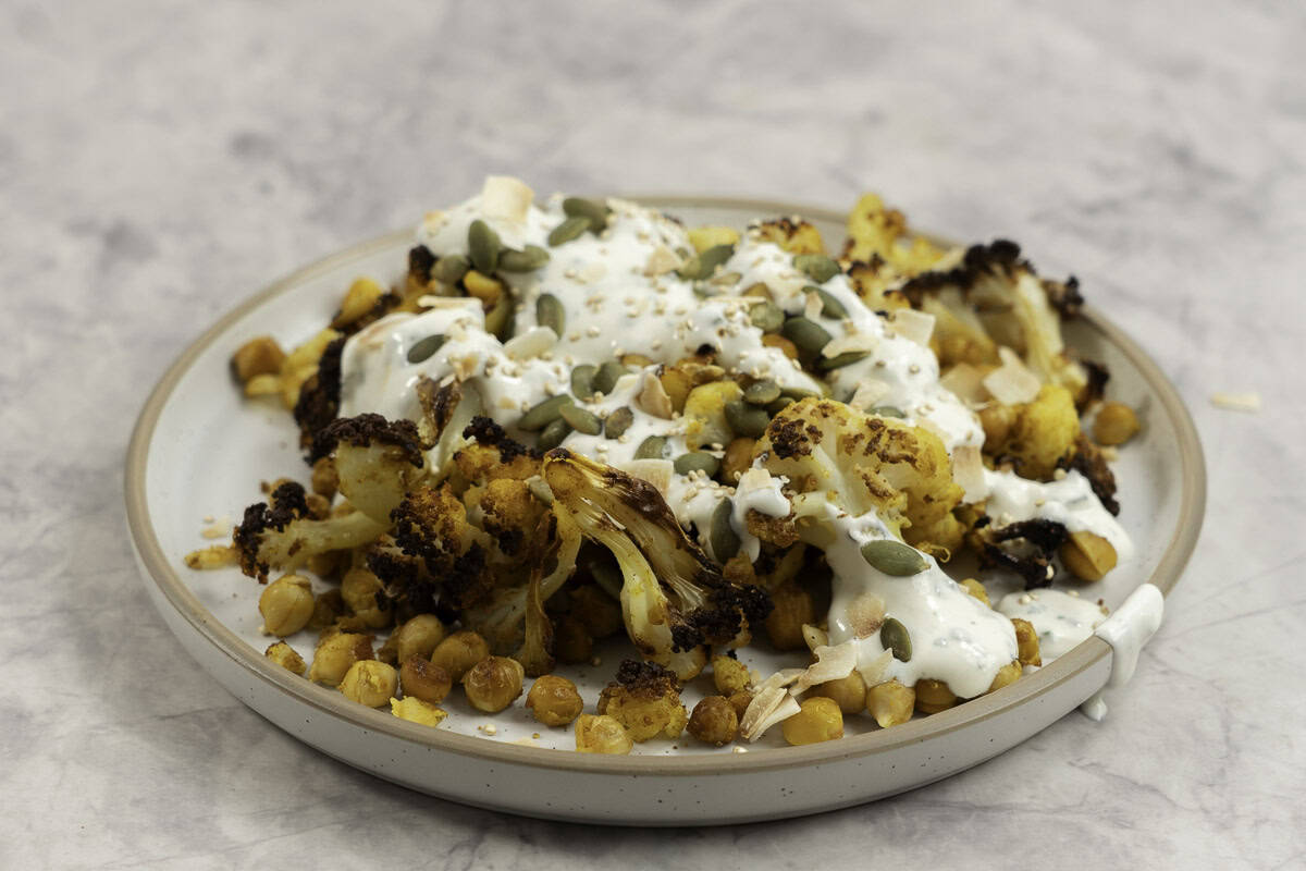 Dinner plate on marble bench top with roasted chickpeas and cauliflower drizzled with taziki dressing and sprinkled pumpkin seeds and coconut flakes.
