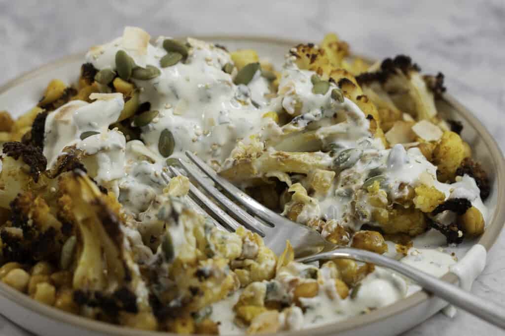 Fork resting on a dinner plate on marble bench top with roasted chickpeas and cauliflower drizzled with taziki dressing and sprinkled pumpkin seeds.