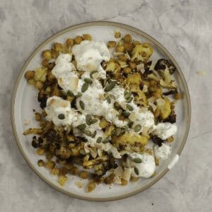 Dinner plate on marble bench top with roasted chickpeas and cauliflower drizzled with taziki dressing and sprinkled pumpkin seeds and coconut flakes.