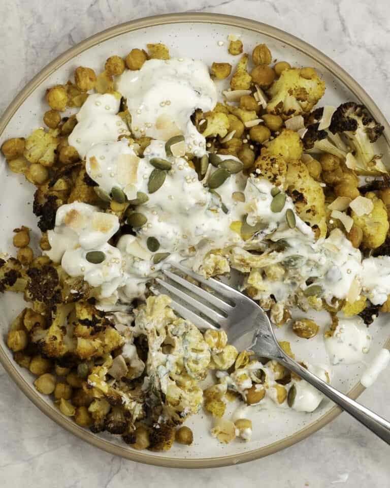 Fork resting on a dinner plate on marble bench top with roasted chickpeas and cauliflower drizzled with taziki dressing and sprinkled pumpkin seeds.