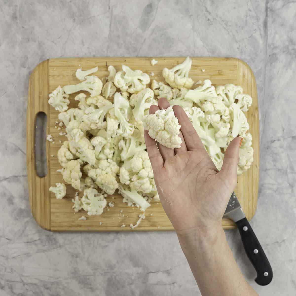 Womans hand holding a cauliflower head above a wooden chopping board of chopped cauliflower.