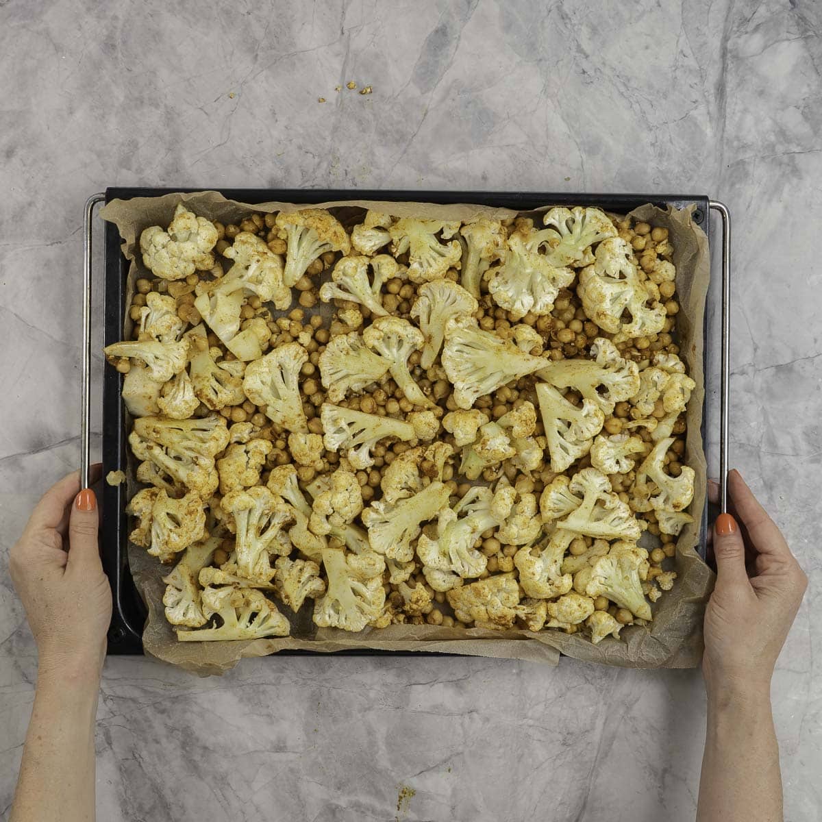Womans hands holding a lined baking tray with cauliflower heads and and chickpeas.