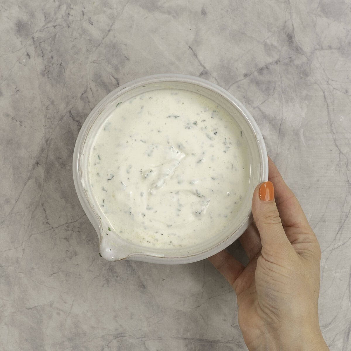 Womans hand holding a bowl of taziki dip on a marble bench top.