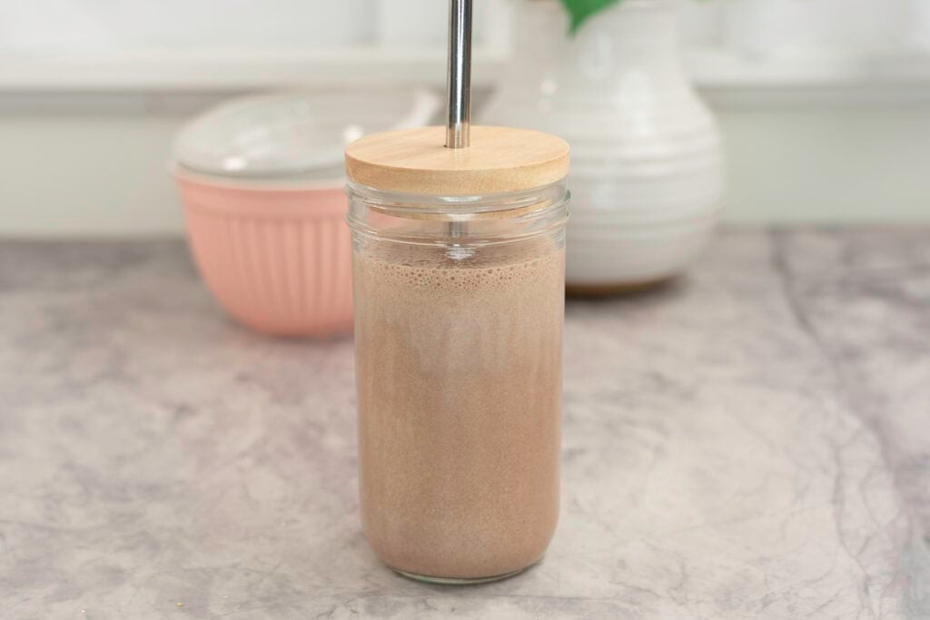 Tall glass with lid and metal straw with chocolate protein shake inside on marble bench top, white ceramic vase and brush pink bowl in background.