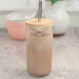 Tall glass with lid and metal straw with chocolate protein shake inside on marble bench top, white ceramic vase and brush pink bowl in background.
