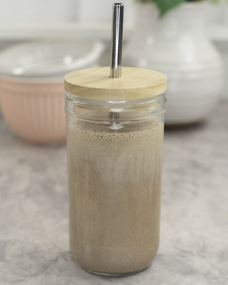 Tall glass with lid and metal straw with chocolate protein shake inside on marble bench top, white ceramic vase and brush pink bowl in background.