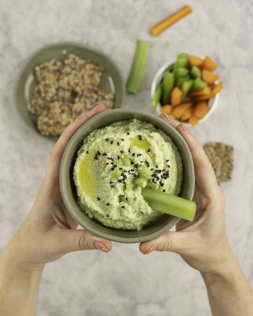 Womans hands holding a small sage green ramekin with homemade hummus inside, topped with edamame and sesame seeds, plate of crackers and sticks of carrot and celery in ramekin in background.