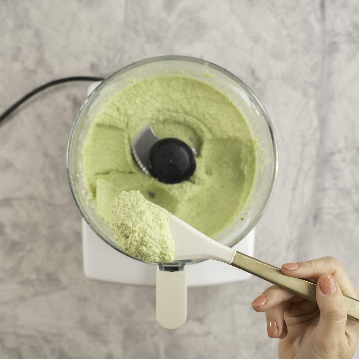 Spatula scooping out hummus from a food processor on bench top.