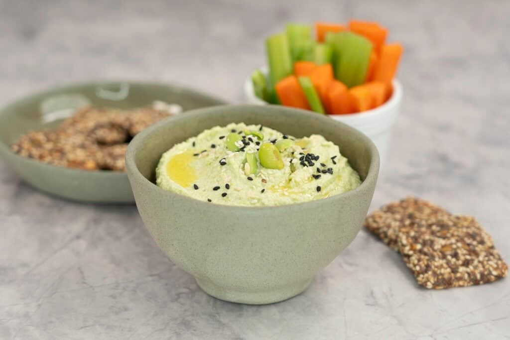 Small sage green ramekin with homemade hummus inside, topped with edamame and sesame seeds, plate of crackers and sticks of carrot and celery in ramekin in background.