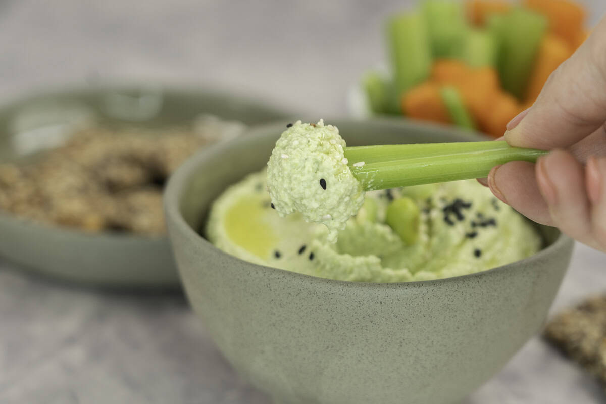Small sage green ramekin with homemade hummus inside, topped with edamame and sesame seeds, woman holding a piece of celery with scoop of hummus ontop and plate of crackers and sticks of carrot and celery in ramekin in background.