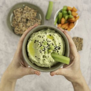 Womans hands holding a small sage green ramekin with homemade hummus inside, topped with edamame and sesame seeds, plate of crackers and sticks of carrot and celery in ramekin in background.