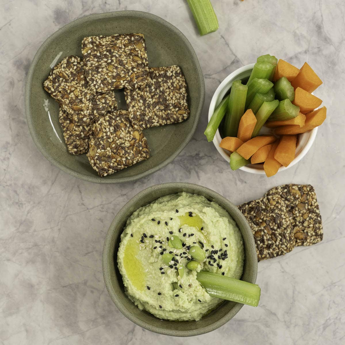 Small sage green ramekin with homemade hummus inside, topped with edamame and sesame seeds, plate of crackers and sticks of carrot and celery in ramekins.