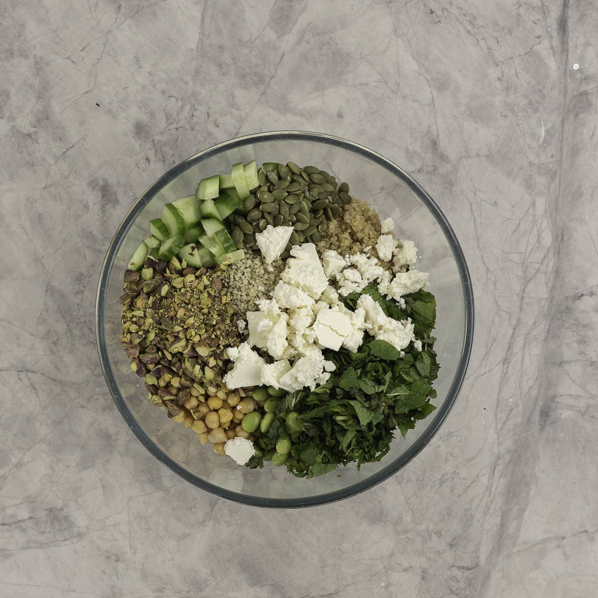 Ingredients for salad inside a large glass bowl on marble bench top. Crumbled feta, disced vegetables, herbs, chickpeas, pistachios and cooked quinoa all visible. 