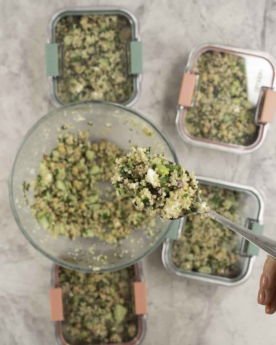 A scoop of salad on a spoon with large glass bowl on bench top below, four containers portioned with salad inside with the lids on.