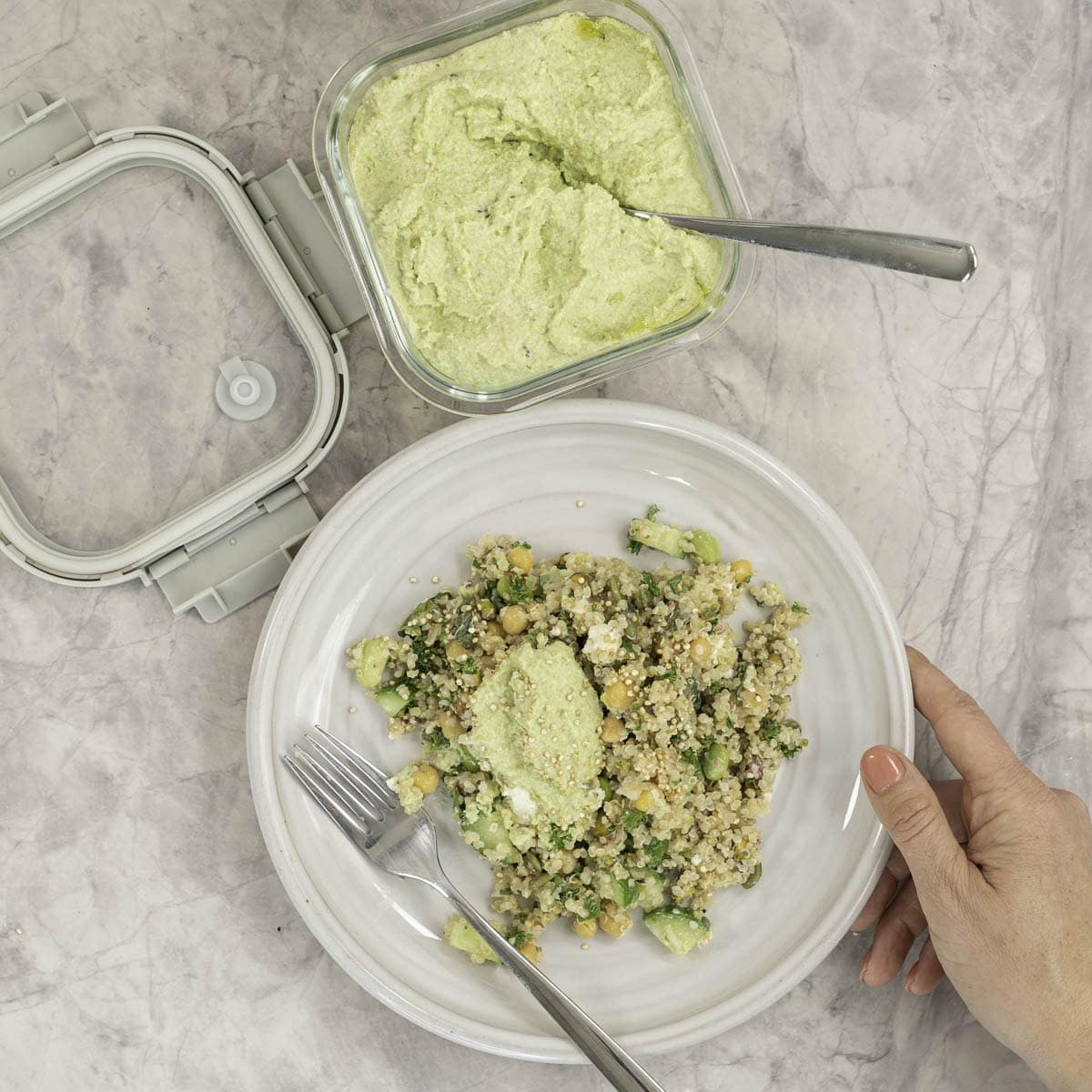 Serving of a salad with dollop of hummus on top with fork and womans hand holding plate on a marble bench top next to a container of homemade hummus.