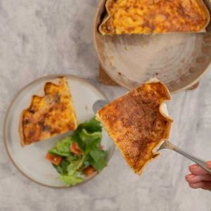 A piece of quiche held up above a dinner plate with quiche and salad, and baking dish with half a quiche inside.