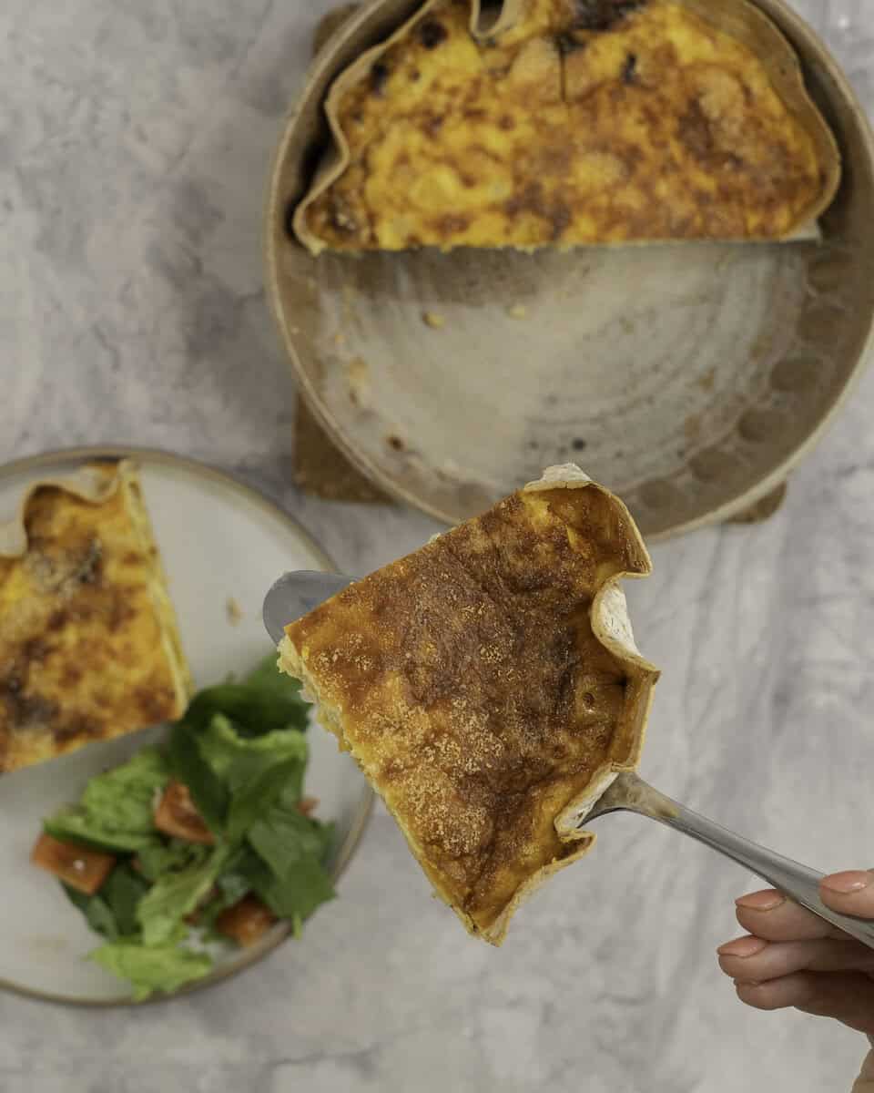 A piece of quiche held up above a dinner plate with quiche and salad, and baking dish with half a quiche inside.