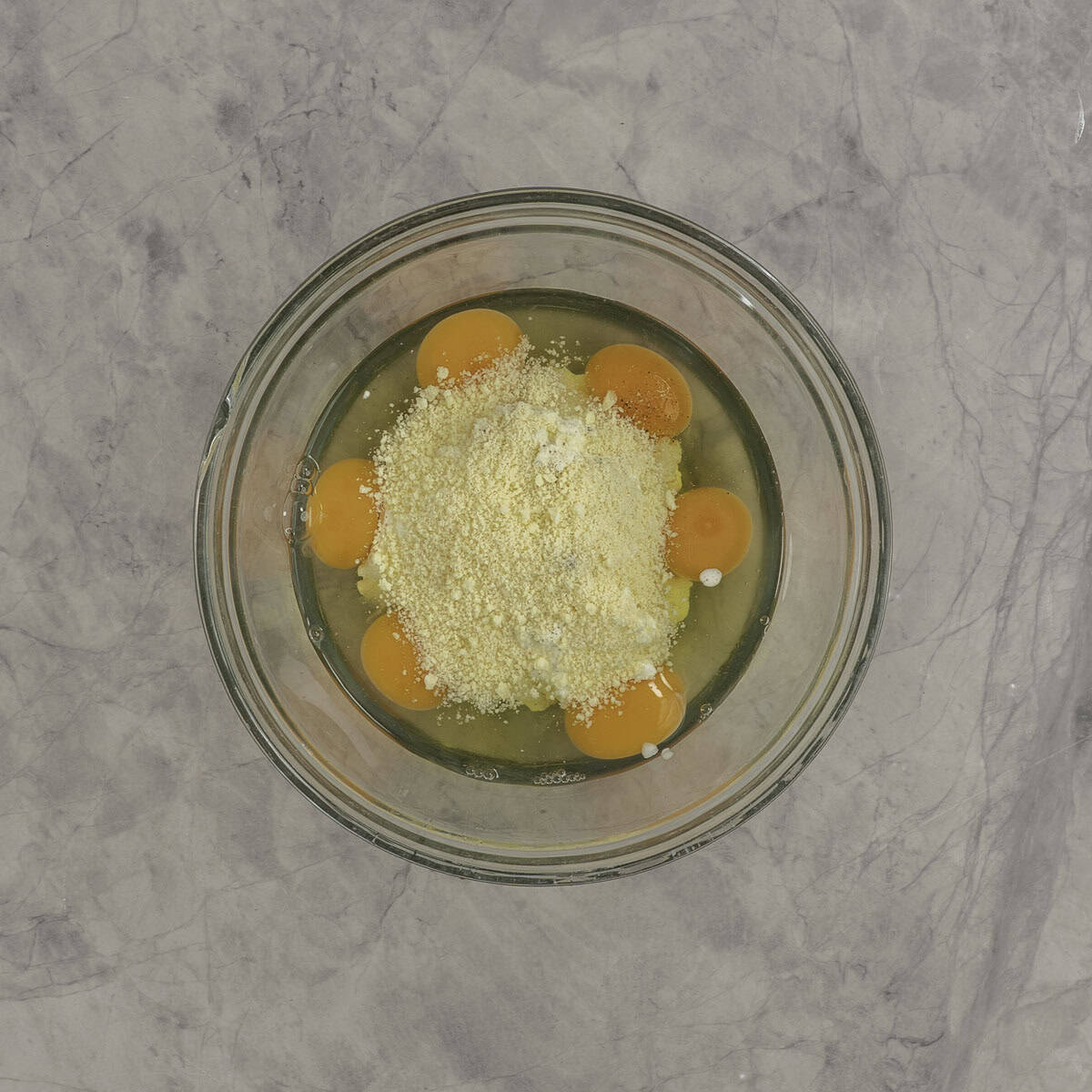 Eggs and parmesan cheese in a large glass bowl on marble bench top.
