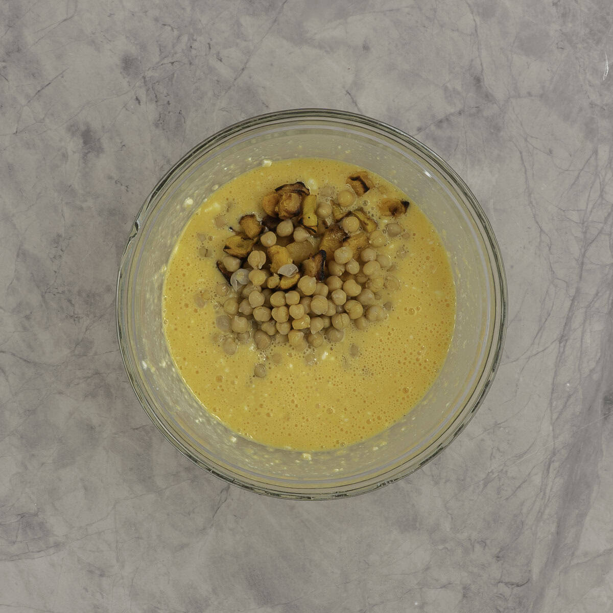Chickpeas, cottage cheese and roasted kumara added into whisked up egg in a large glass bowl on marble benchtop.