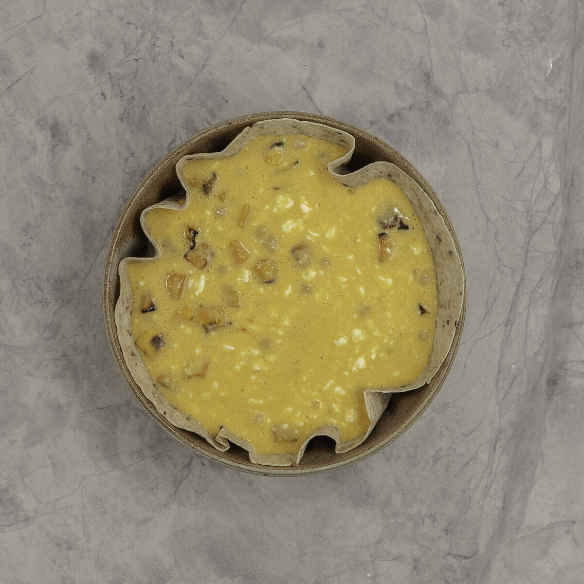 Uncooked quiche with a tortilla base in a round baking dish on marble benchtop.