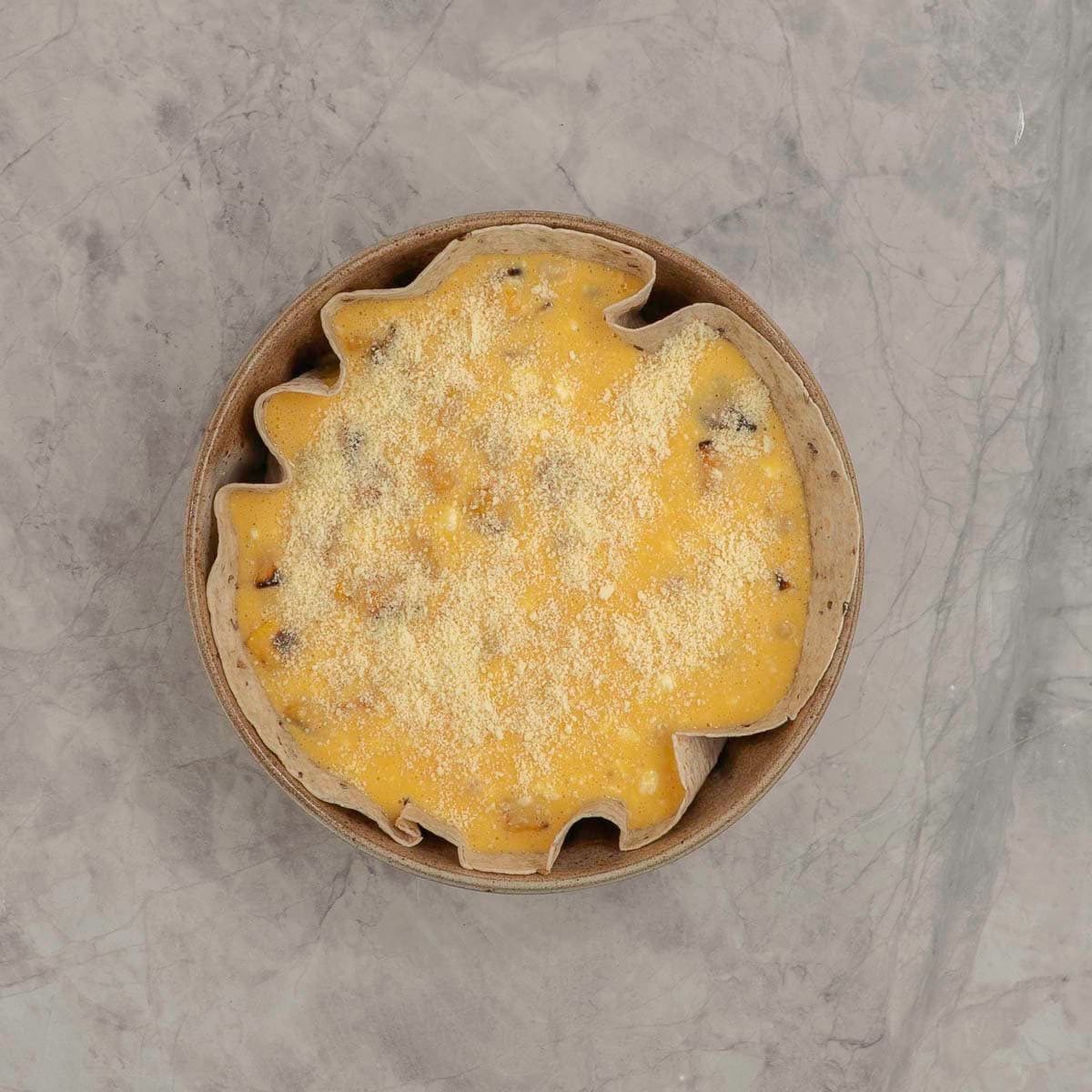 Uncooked quiche topped with parmesan with a tortilla base in a round baking dish on marble benchtop.