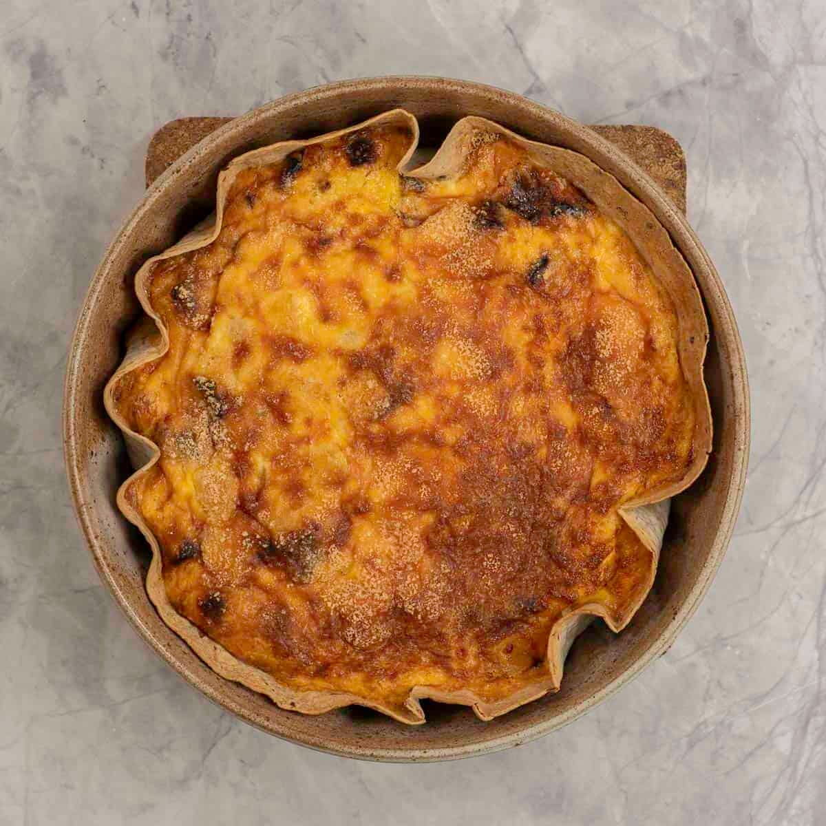 A quiche golden brown on the top with tortilla base in a round baking dish on marble benchtop.