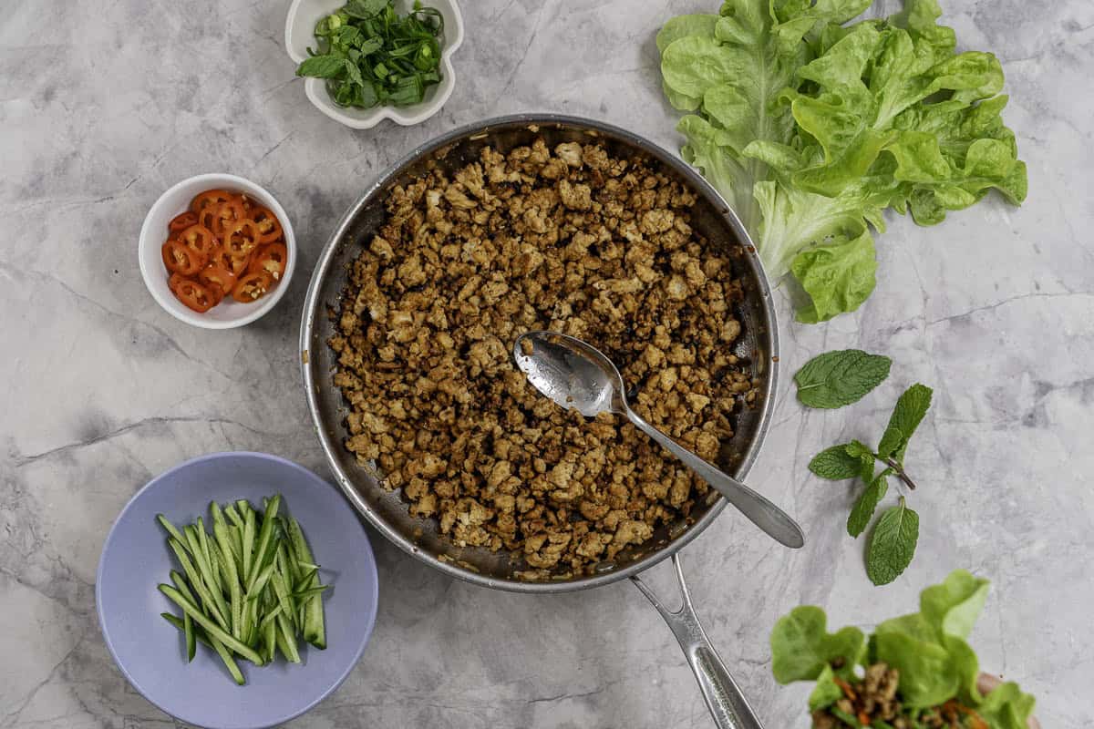 Chicken mince in a pan on benchtop with plates of thinly sliced cucumber, sliced chilli, spring onion and fresh lettuce.