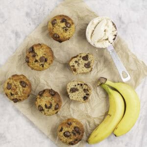 Baking paper on marble bench top with 5 muffins, one cut in half laying down showing fluffy inside and melted chocolate chips, two bananas and a measuring cup of protein powder.