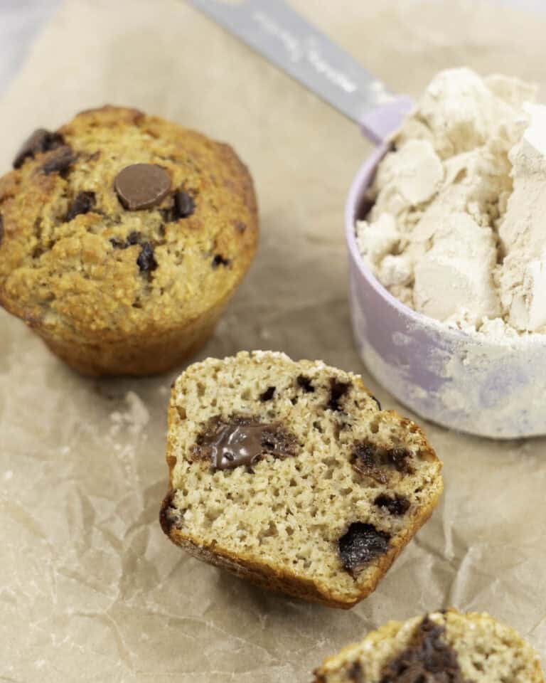 Baking paper on marble bench top with 5 muffins, one cut in half laying down showing fluffy inside and melted chocolate chips next to a measuring cup of protein powder.