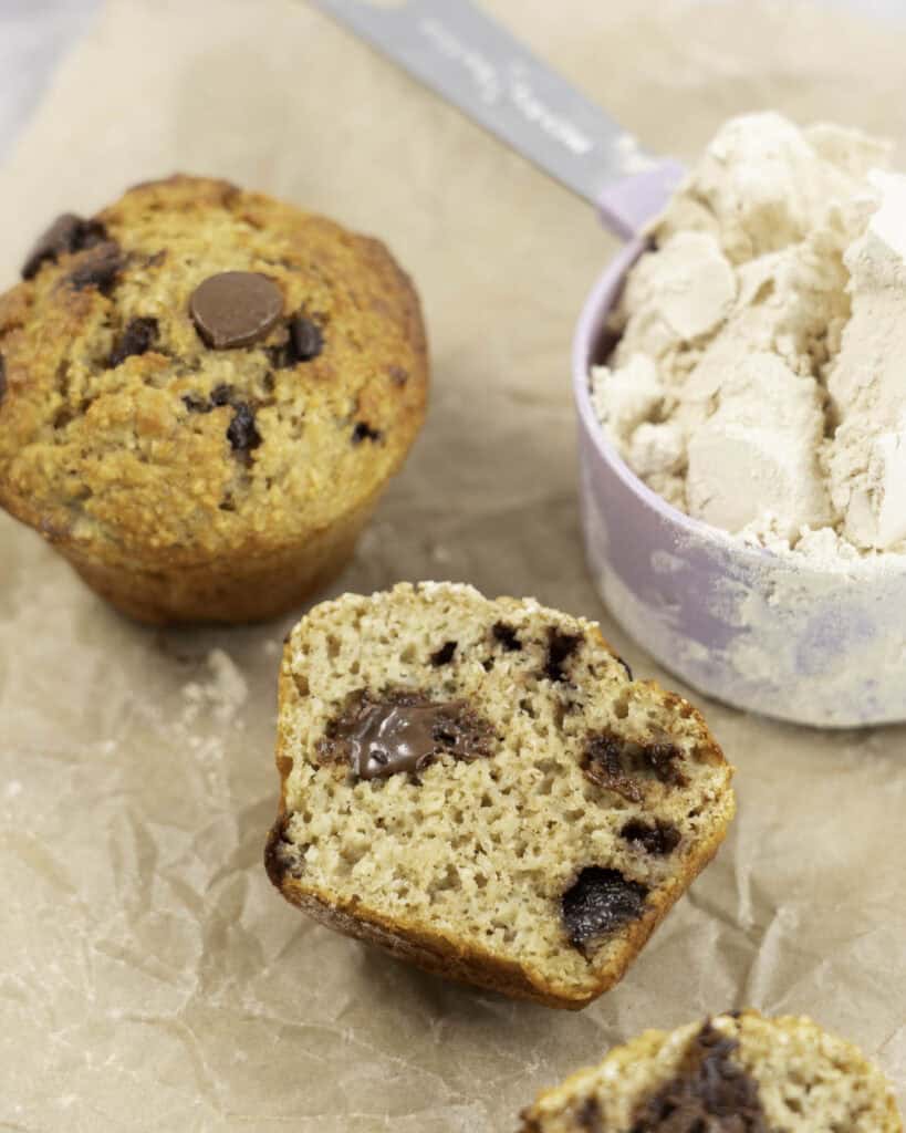 Baking paper on marble bench top with 5 muffins, one cut in half laying down showing fluffy inside and melted chocolate chips next to a measuring cup of protein powder.