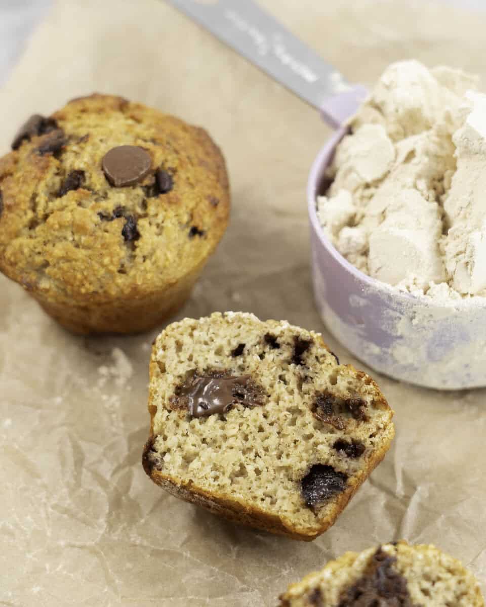 Baking paper on marble bench top with 5 muffins, one cut in half laying down showing fluffy inside and melted chocolate chips next to a measuring cup of protein powder.