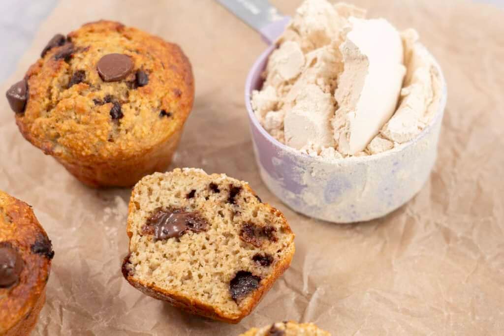 Baking paper on marble bench top with 5 muffins, one cut in half laying down showing fluffy inside and melted chocolate chips next to a measuring cup of protein powder.