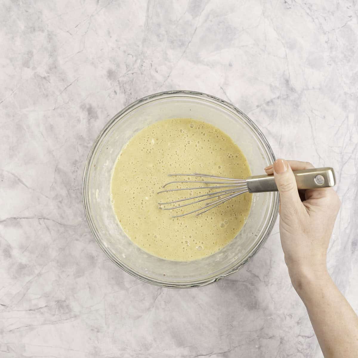 Womans hand holding a whisk stirring wet ingredients in a glass bowl on benchtop.