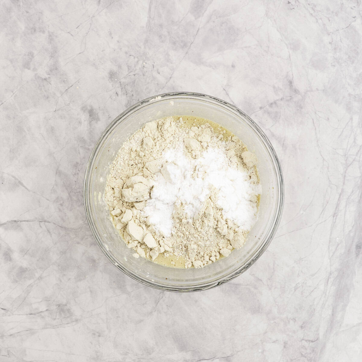 Dry ingredients added on top of wet mixture in glass bowl on marble benchtop.