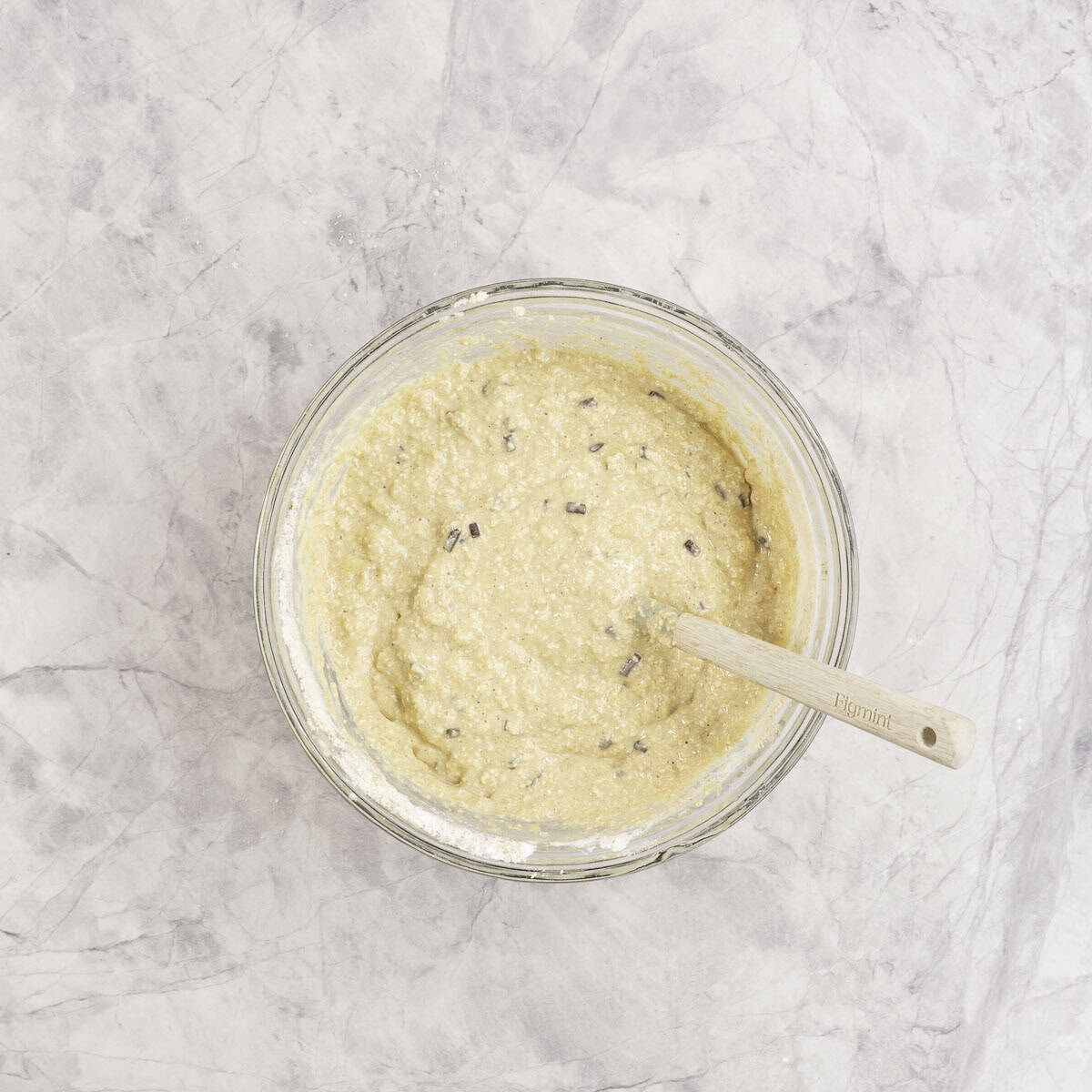 Mixture for muffins in glass bowl with spatula inside on marble bench top.