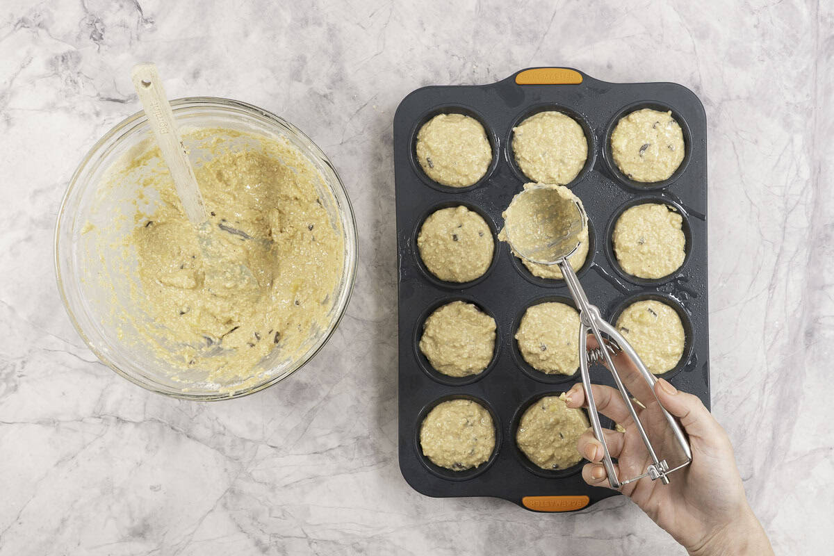Womans hand holding a cookie scooper with muffin batter scooped into 12 tray muffin tin below, residue of batter in glass bowl on benchtop.