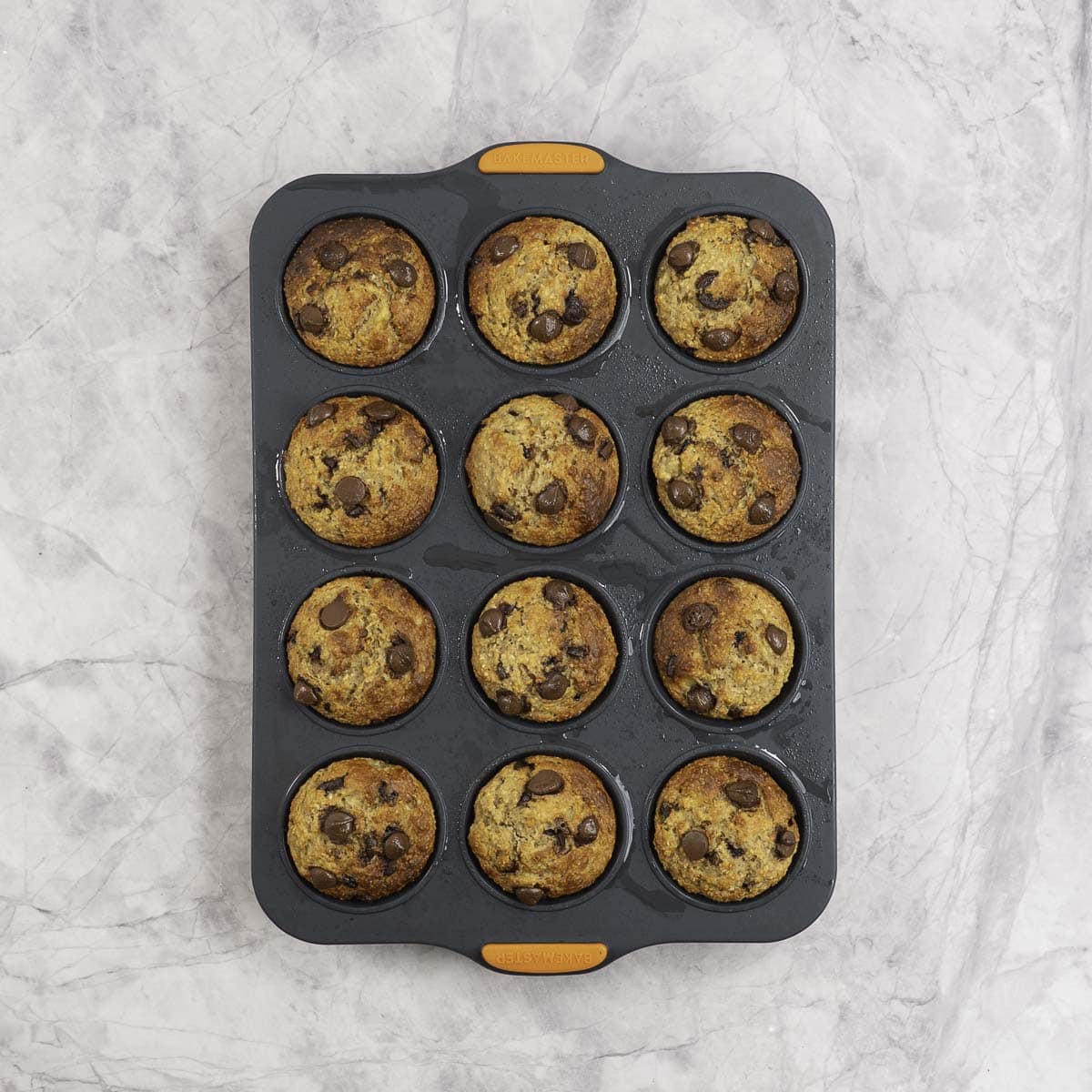 Golden brown muffins in a 12 tray muffin tin on marble benchtop.